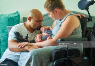 Disabled mother sitting in her wheelchair in front of newborn baby's crib holding her newborn son, husband is kneeling beside her.